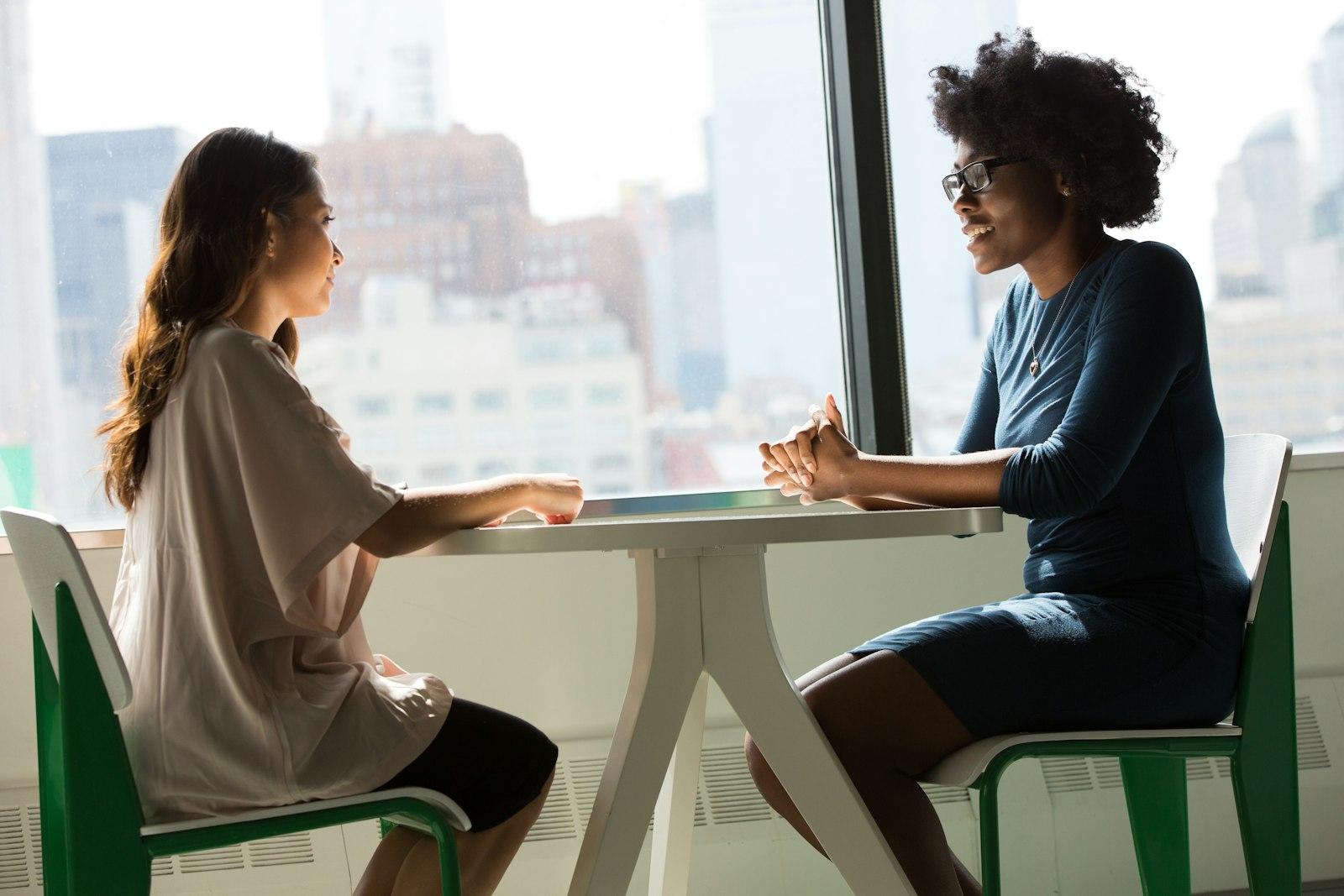 Two women sitting across a table in conversation by a window